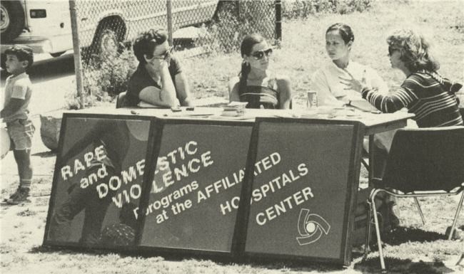 four women sitting behind a table with a sign "Rape and Domestic Violence Programs at the Affiliated Hospitals Center"